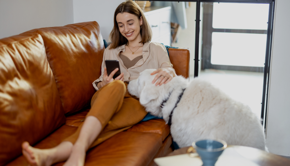 Woman smiling on the sofa, looking at her phone while stroking her dog