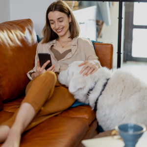 Woman smiling on the sofa, looking at her phone while stroking her dog