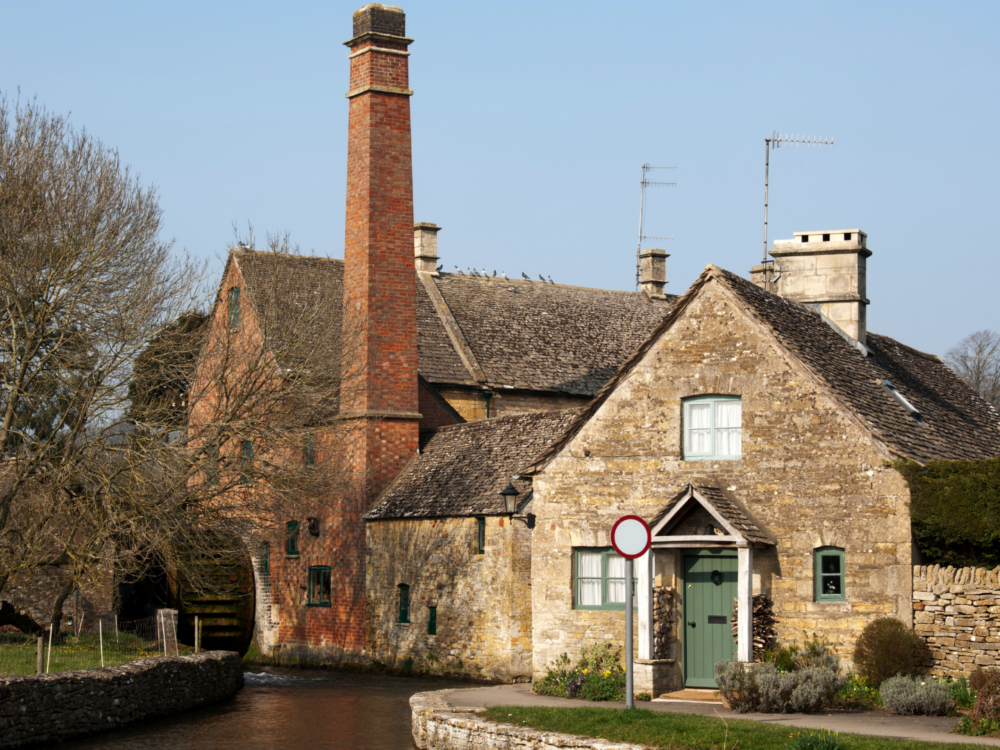 A traditional Grade II-listed stone cottage in the Cotswolds, with a watermill and chimney visible beside a tranquil stream. The scene evokes rural charm and architectural heritage in a highly sought-after countryside setting.