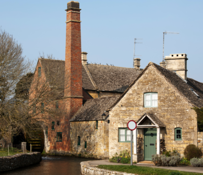 A traditional Grade II-listed stone cottage in the Cotswolds, with a watermill and chimney visible beside a tranquil stream. The scene evokes rural charm and architectural heritage in a highly sought-after countryside setting.
