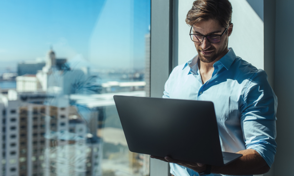 A professional man stands by a window in a high-rise building, looking at his laptop with a focused expression. The city skyline is visible in the background, symbolising global connectivity and cross-border investment opportunities.