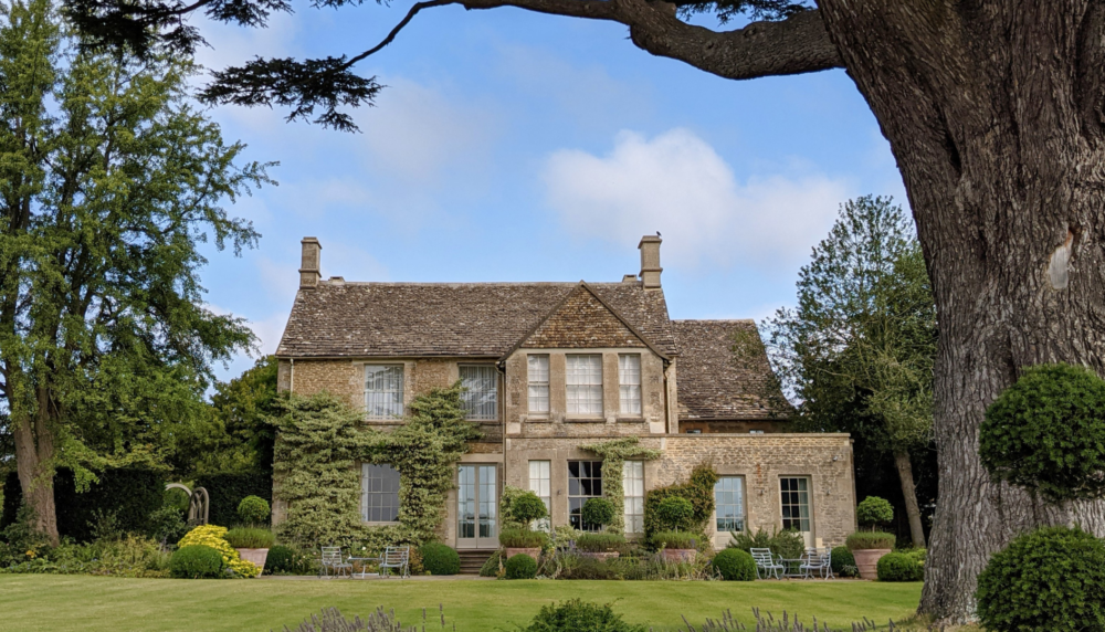 A grand English country estate surrounded by mature trees and manicured gardens, viewed from a distance under a clear sky. The historic architecture and expansive grounds reflect the scale and character of a high-value rural property.