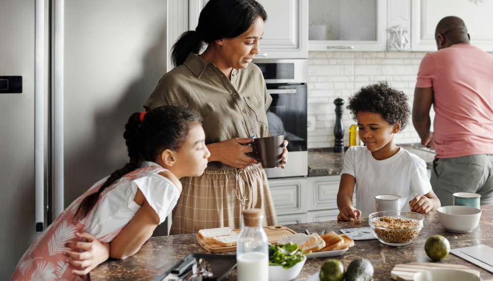 A family gathered in a bright, modern kitchen during breakfast. A mother holds a coffee mug while talking to her children at the counter, where cereal, toast and fresh ingredients are laid out. A father prepares something in the background, symbolising a warm, secure family routine.