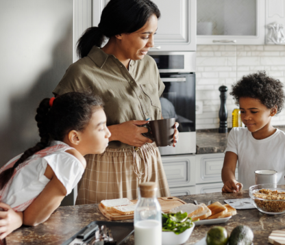 A family gathered in a bright, modern kitchen during breakfast. A mother holds a coffee mug while talking to her children at the counter, where cereal, toast and fresh ingredients are laid out. A father prepares something in the background, symbolising a warm, secure family routine.