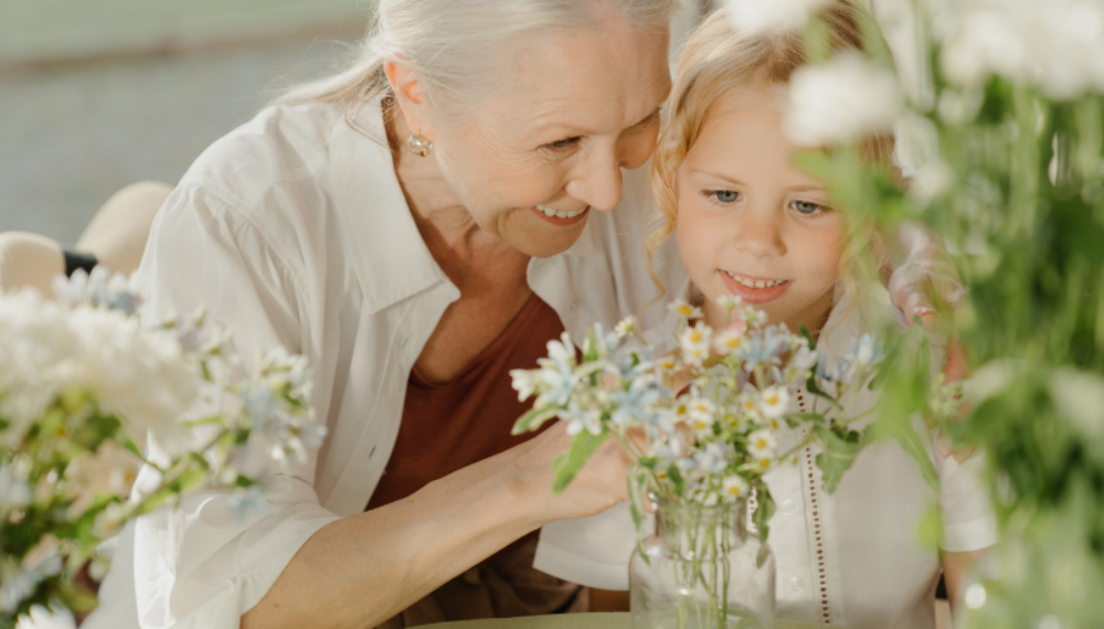 Elderly woman and young girl smiling together while arranging flowers, symbolising intergenerational support and family connection.