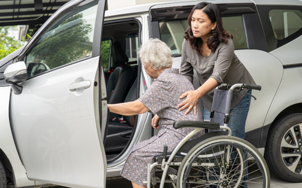 Adult daughter helping her elderly mother in a wheelchair into a car, representing family caregiving and financial support in later life.